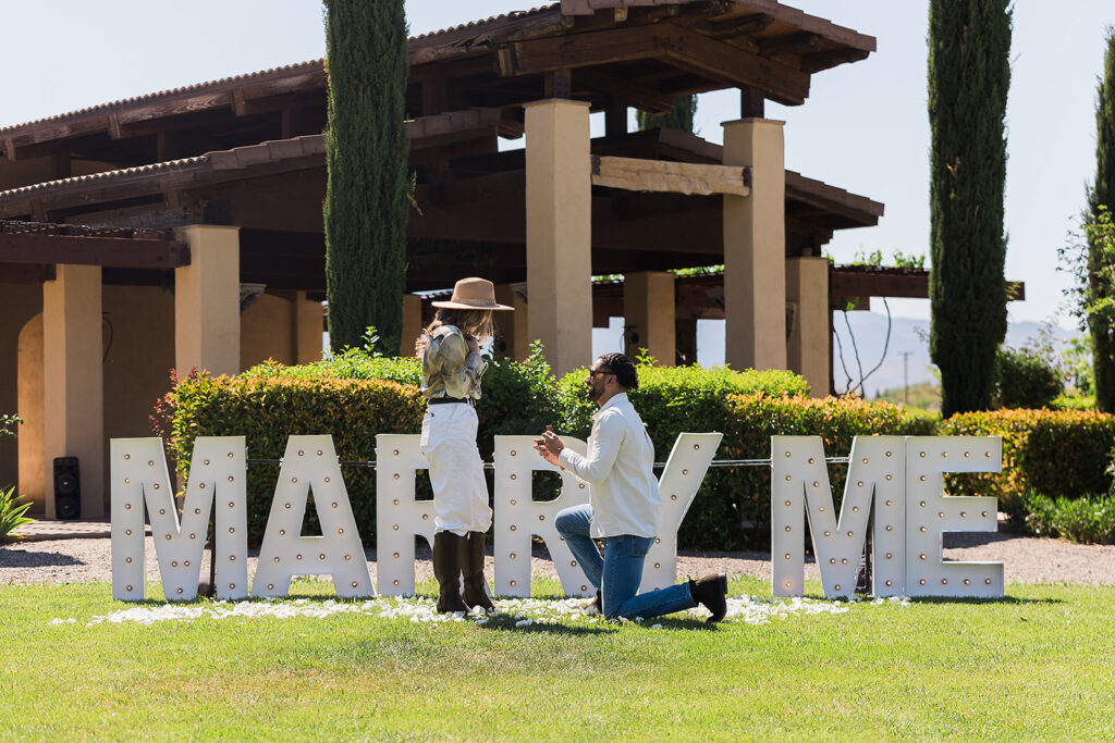 groom down on one knee proposing in front of marry me marquee