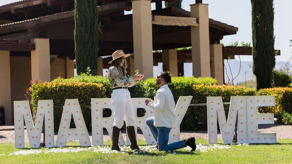 groom down on one knee proposing in front of marry me marquee