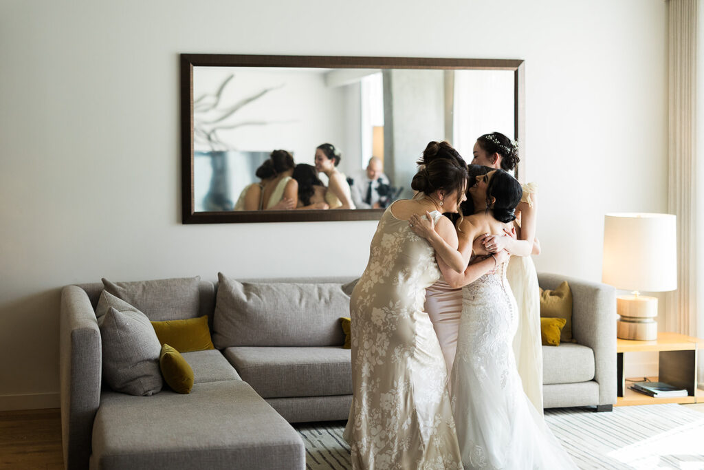 bridal party hugs bride in hotel room during first look of bride in gown