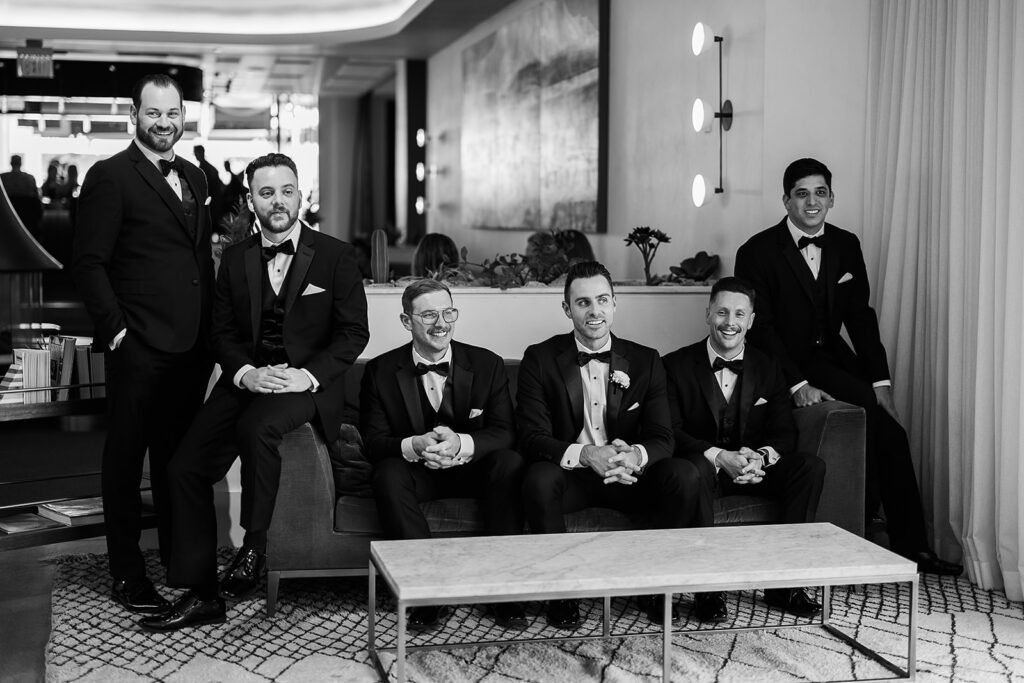 groomsmen sit on couch in suits and sunglasses in hotel